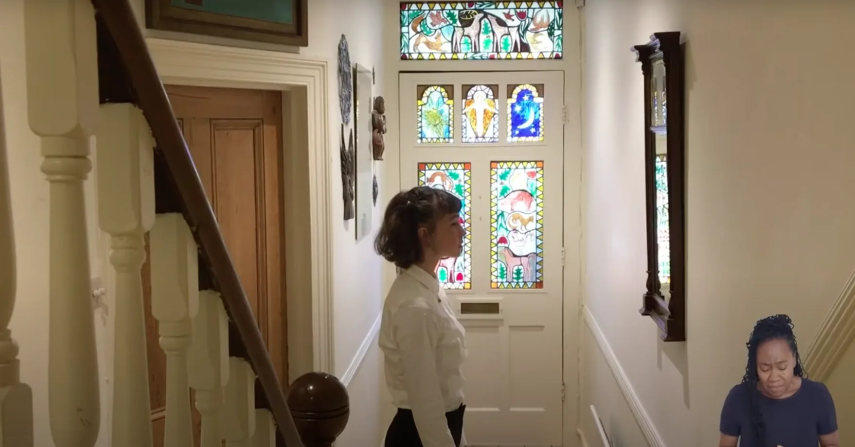 A screenshot from our film 'What Do You See In Me'. Tilly stands in the hallway of her family home, a front door inlaid with colourful stained glass behind her. Jacqui, a sign language interpreter, can be seen in the bottom right of the screen