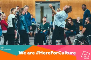 A group of people stand around laughing in the rehearsal room, with a banner beneath which reads we are here for culture