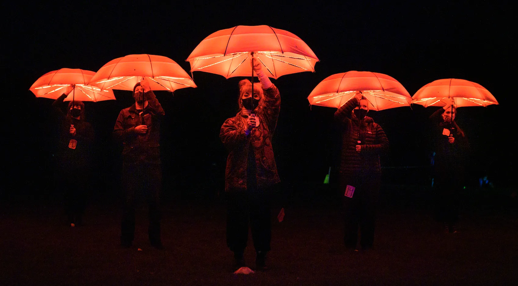 Performers stand under the canopy of red lit LED umbrellas. They are wearing COVID masks that cover their mouths and noses.