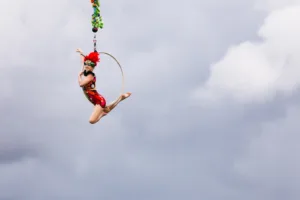 An aerialist dressed in a pretty red costume and feathered headpiece hangs in the air on her hoop against a cloudy background