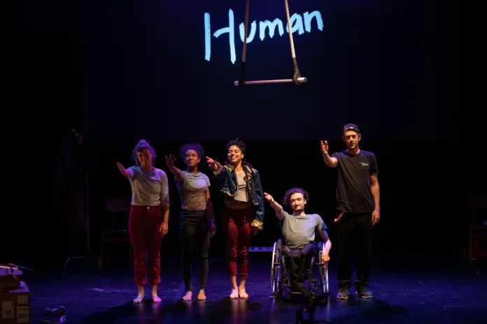 Four performers stand looking at their audience, with their arms outstretched. Behind them the word Human is displayed on a screen, with a hanging trapeze in front of it