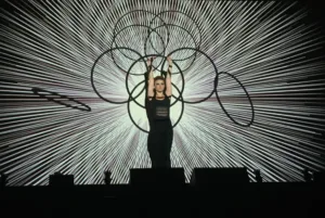 A Hula Hooper stands in front of a video mapped screen, holding her hula hoops high above her head