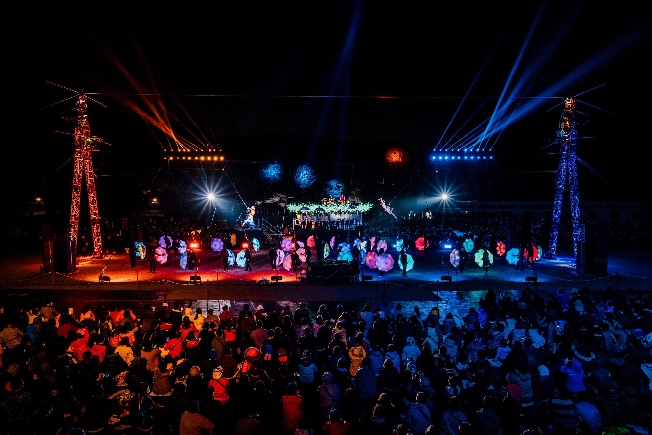 A large audience look onto a stage filled with bright LED coloured umbrellas being manipulated by onstage performers. There are two aerialists hanging from the big metal structure and both are lit by a bright white light behind them.