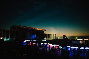 A trail of LED umbrellas light up the night sky, illuminating a path to a main stage at a festival