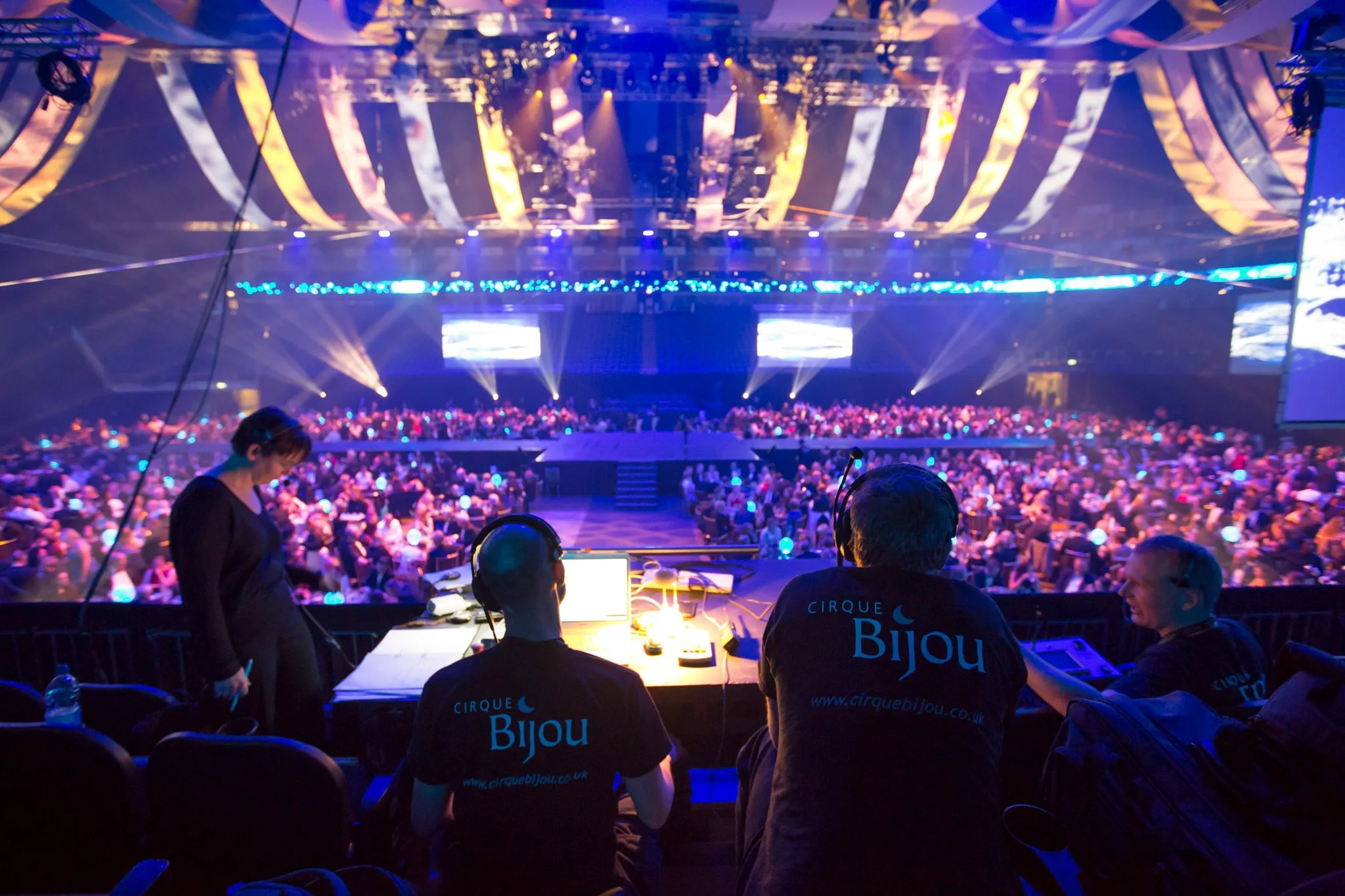 In the foreground are three members of crew, looking out over a lit stage and audience, waiting for a production to begin