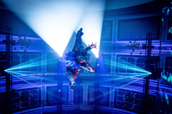 A performer hands suspended from the ceiling, reaching down and towards the camera. Blue lights and lasers surround her