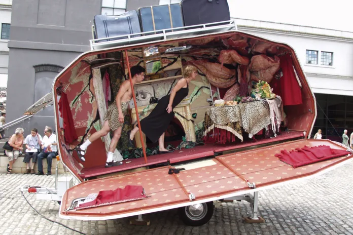 Bristol Harbour Festival. Two performers, 1 female and 1 male, lean forwards in an elegantly decorated caravan, with suitcases on a rack above. There is a pole in front of them and a decorated leopard print table up the other end of the vehicle.