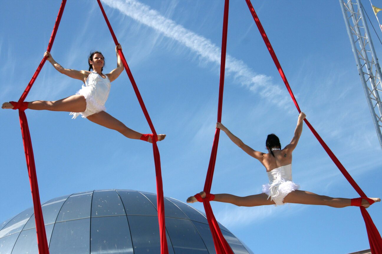 Bristol Harbour Festival, 2008. Two female performers hold the splits in red silks. They are wearing white leotards and the performer on the left is facing forwards whilst the other performer faces the back. There is a blue sky behind them.