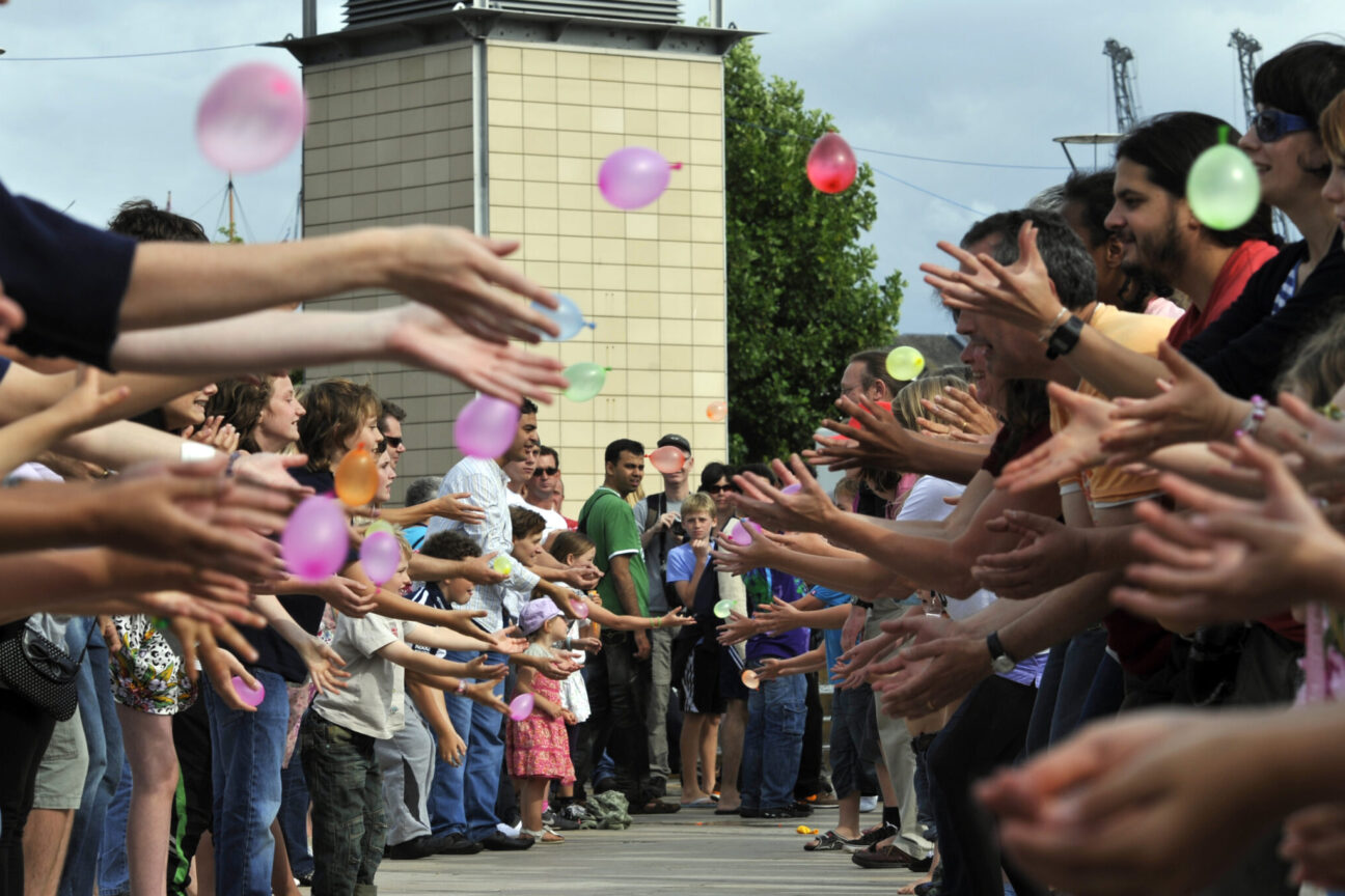 Balloon toss at Bristol Harbour Festival, 2009. Children and adults are gathered in two lines facing into each other, there are colourful balloons being thrown ,and already thrown, towards to opposing line. There are happy and fearful expressions on the faces of participants.