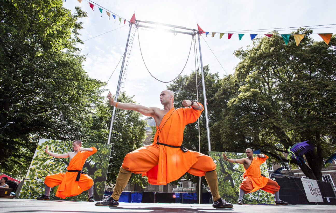 Bristol Harbour Festival, 3 performers stand in orange costumes holding a martial arts pose, their arms out to the right. They are stood on the Cirque Bijou stage with a rig above them.