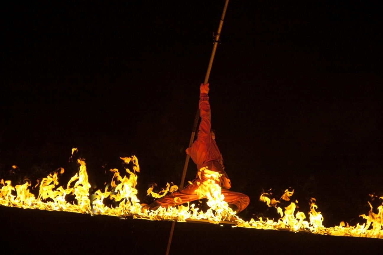 A fire high wire walker performers on a flaming wire, balance poles extended, against a black night sky. Goodwood FOS 2015