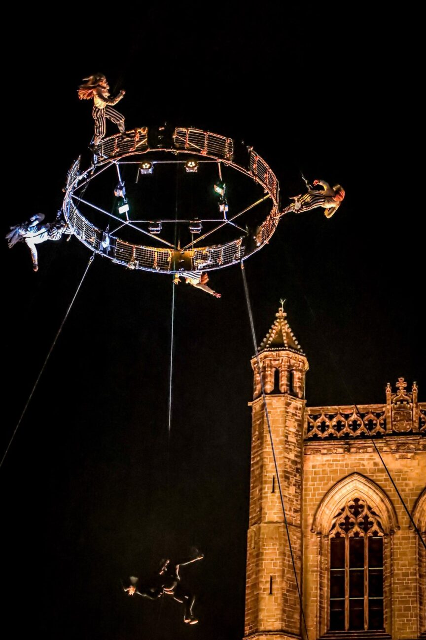 Paisley Halloween Festival. Four performers in black and white striped trousers and white tops are running around an Aerial carousel suspended in the air by a crane.