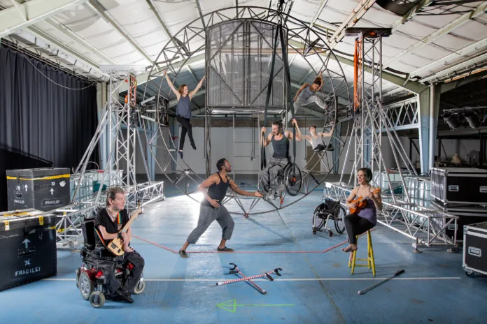 A group of disabled and nondisabled performers stand infront of a large piece of metal circus equipment