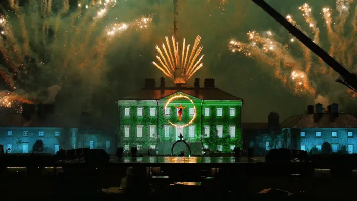 A stately home is illuminated green with projections, with fireworks going off all around it. In the foreground and aerialist performers in a hoop of fire