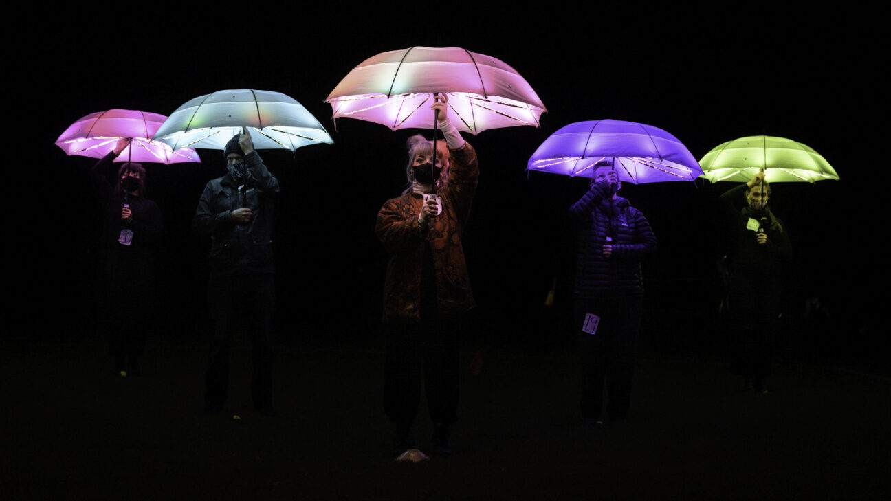 5 performers with face masks hold different coloured LED umbrellas above their heads.