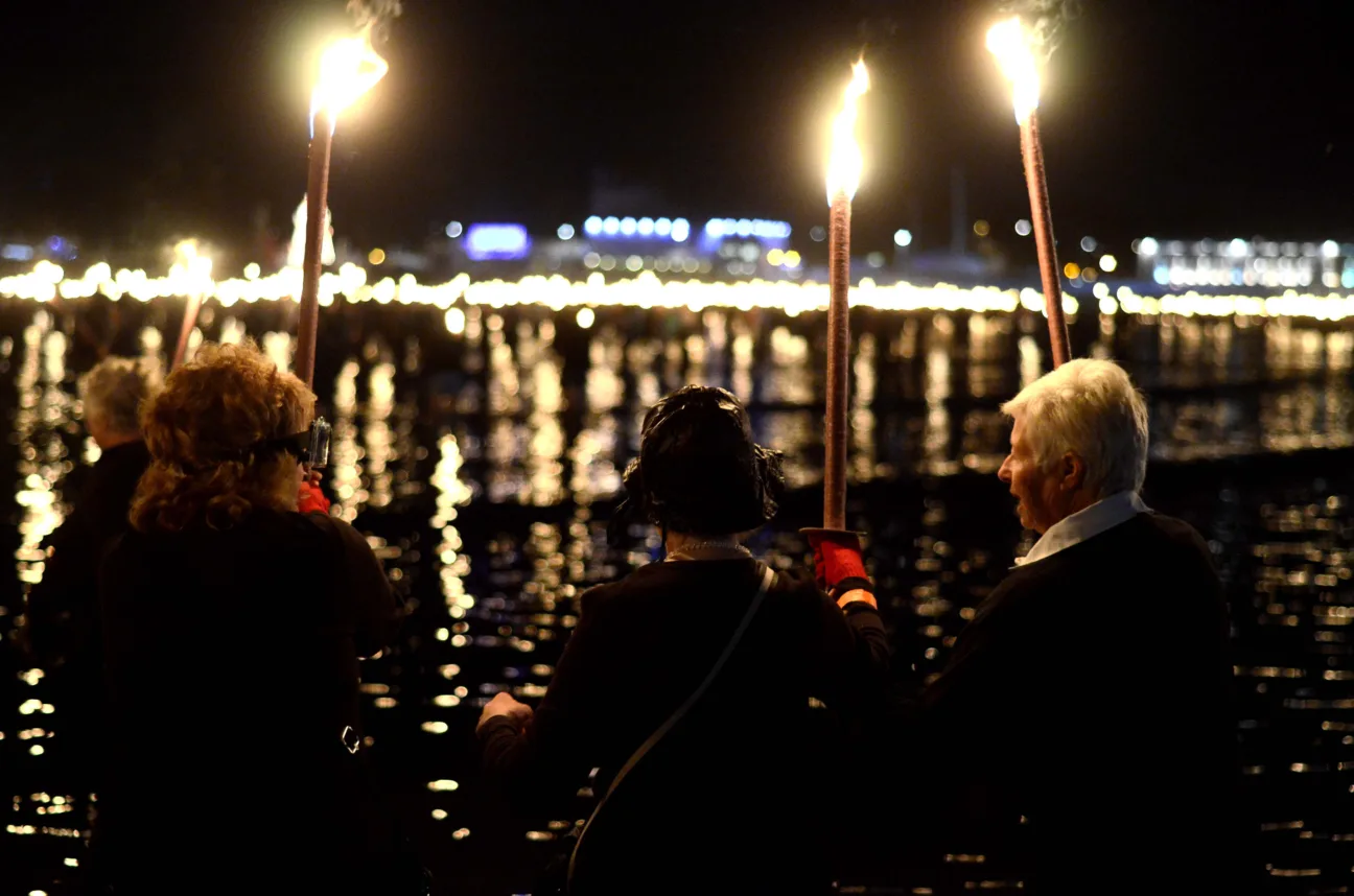 3 people, 2 female and 1 male, stand facing away from the camera holding up fire torches. In front of them is water.