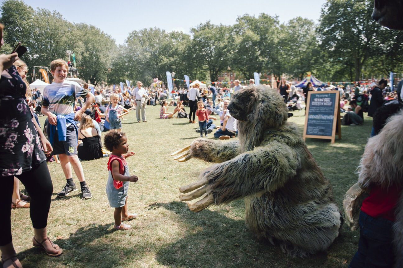 Bristol Harbour Festival. A little girl is facing a performer in a giant sloth costume who opens their arms to offer a hug. In the background there are banners and marques as well as many children and adults at the festival.