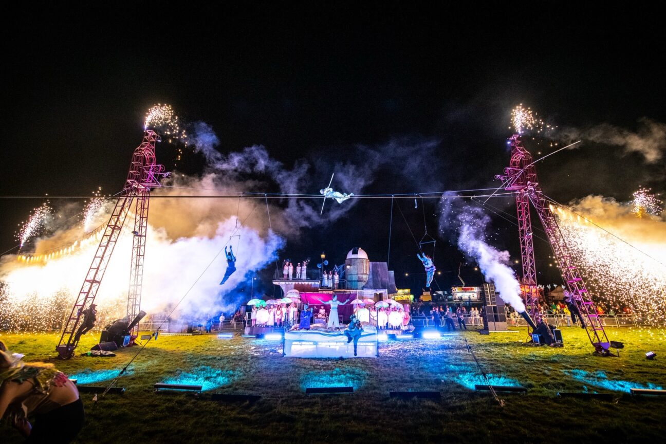 Camp Bestival, finale show. Off of a large pink structure with fire coming out of it, there are performers: one in the centre on a tight wire, two trapeze artists hanging either side below them. There is a lady sat on a box centre stage on the floor, playing the violin, and a man to her left also playing violin. There is a performer dressed in white in the middle of them with their arms out to the side. Behind them there's a procession of people lined up in white with LED umbrellas behind the metal structure.