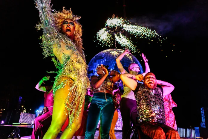 Camp Bestival. Performers are partying and dancing infront of a giant disco bool and bright pyrotechnics in colourful disco outfits with their arms proudly out.