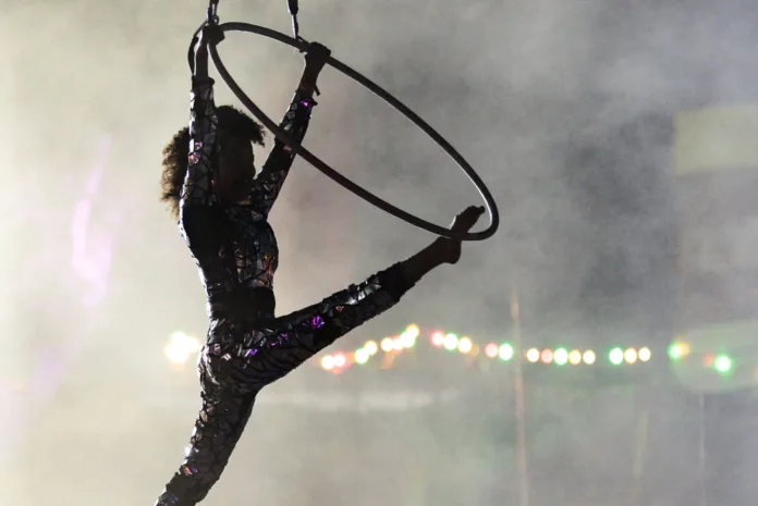 Camp Bestival, female performer performers a split hold, one leg on the hoop, one straight back out the hoop. Behind her is a smoky, blurred background.