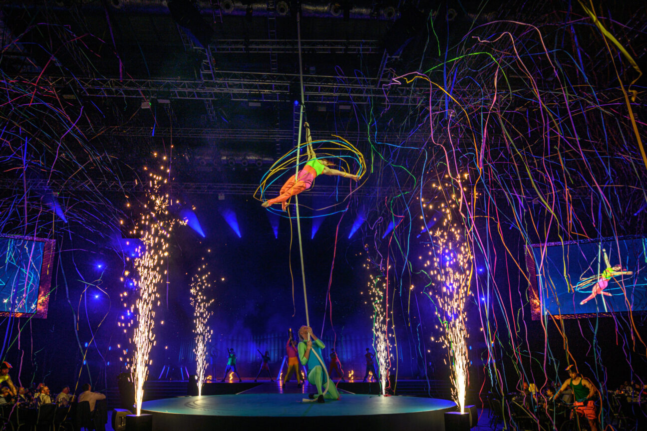 On a circular stage an aerialist hangs from a strap mid-air waving neon coloured ribbons, she is held up there by another performer at the bass of the stage. Simultaneously, there are pyrotechnics and strings of neon confetti released. The performers are wearing neon costumes.