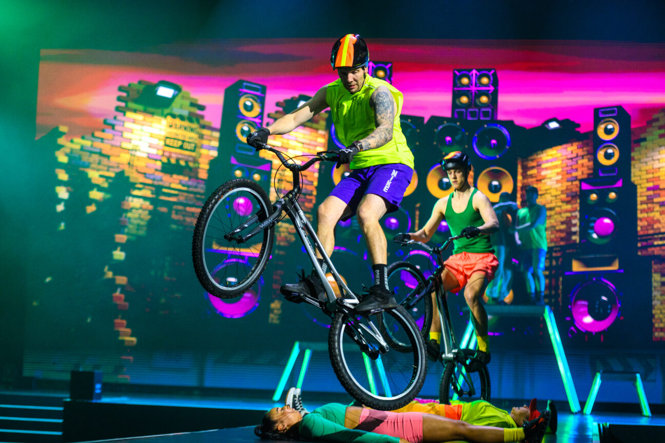 In front of a projection of colourful speakers. Cyclist stunt performers air captured mid-air over 2 3 performers led in a line on the floor. All performers are wearing neon clothing in a variety of colours.