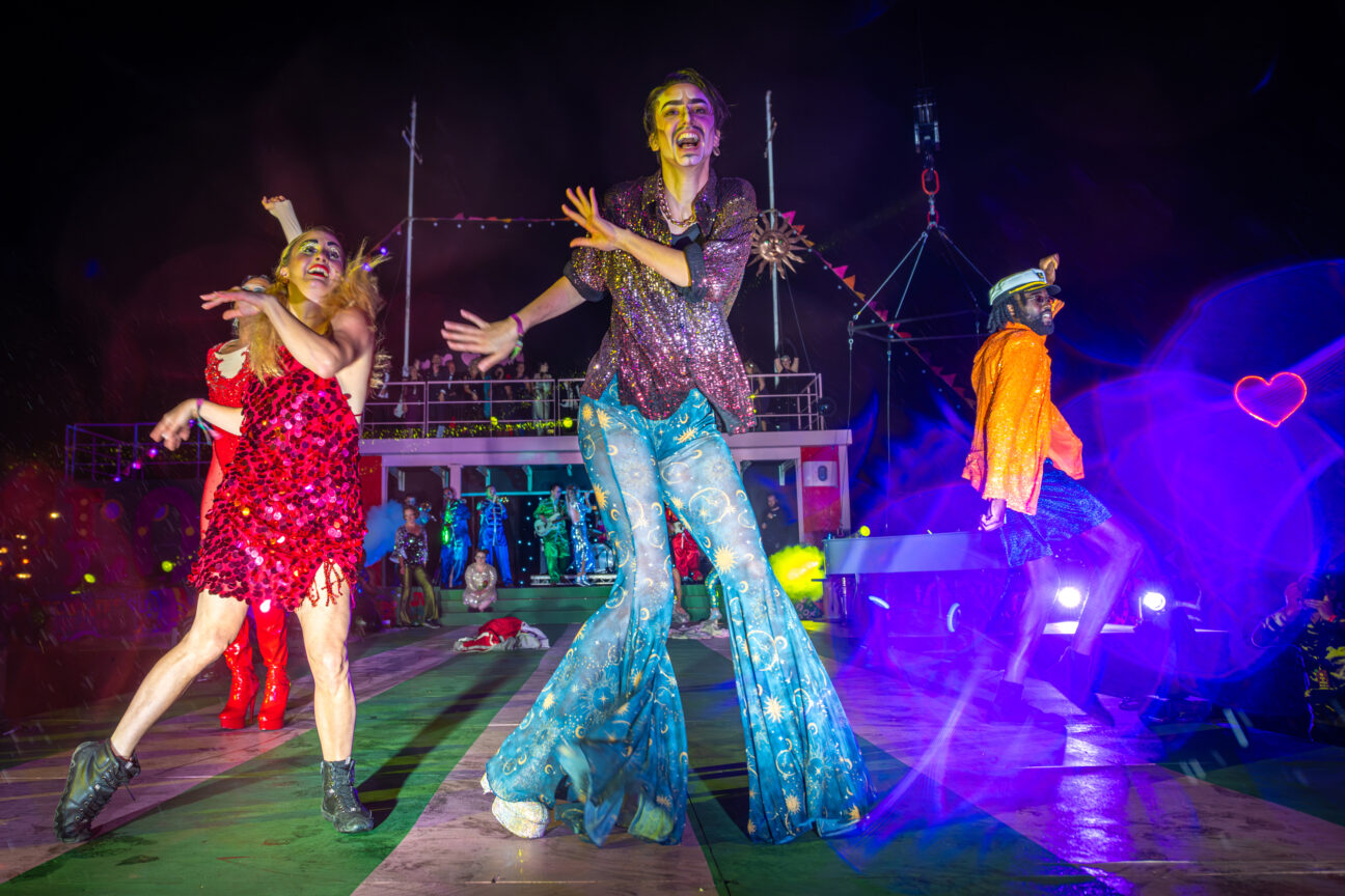 Performers on stage at Camp Bestival, Dorset. Wearing funky disco clothes, flares and sparkly dresses they are twisting their legs as a dance move.