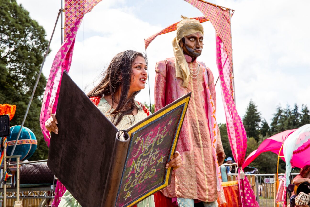 A female performer holds a large prop Storytelling book which says "Night Night Tales". She is reading it to the audience. There is a statue to her right.