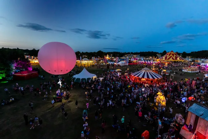 Camp Bestival Birdseye view of an aerial performer attached to a large pink ballon. There are big tops in the background and large crowds gathered around the big tops.