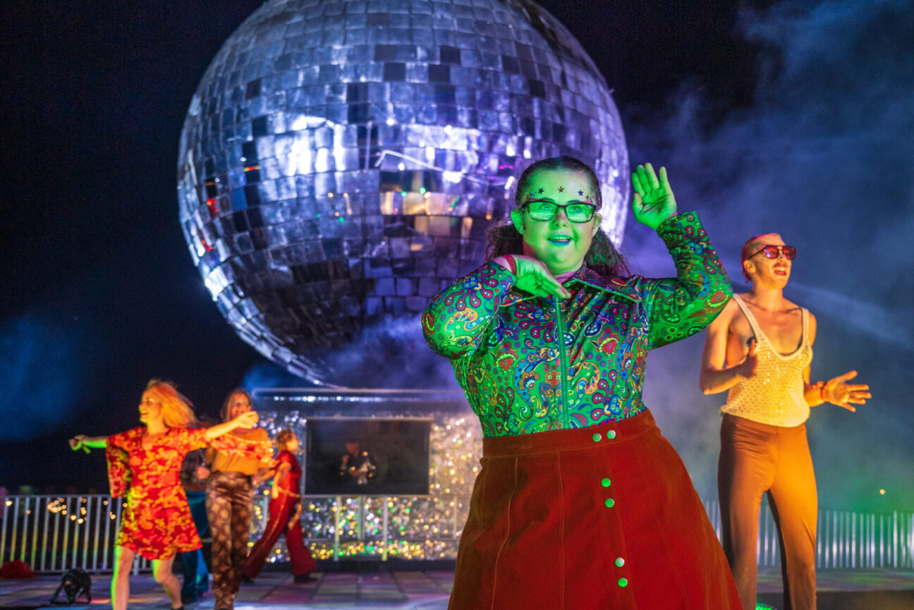 Performers in bright disco clothing stand in front of the giant silver disco ball at Camp bestival.