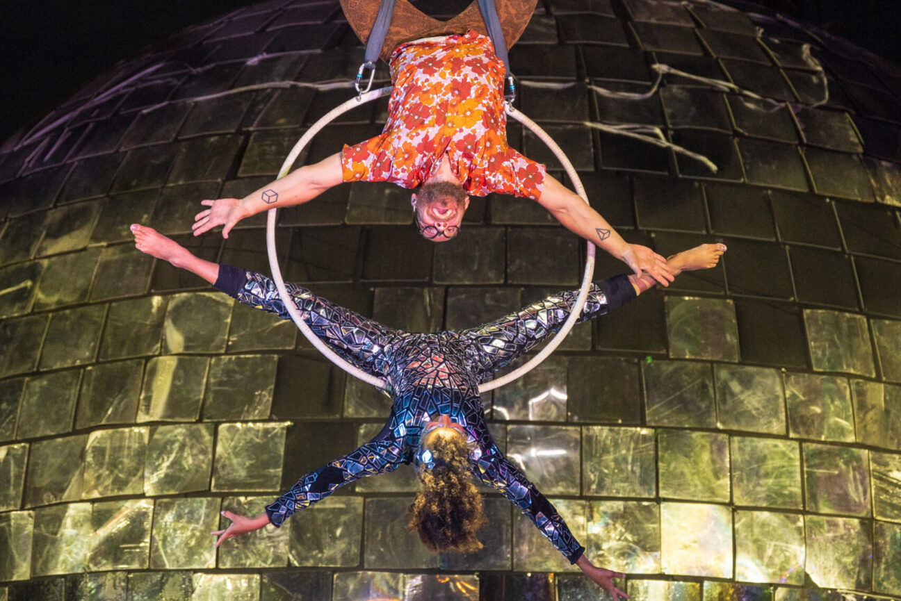 Two aerial performers hang above and below the hoop in front of the disco ball in flowery and shiny costumes.