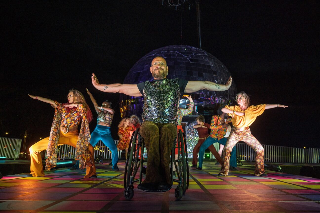 7 performers in colourful disco outfits hold poses with their legs bent and arms out in different directions. The centre male performer uses a wheelchair and has his arms out fiercely with a large smile.