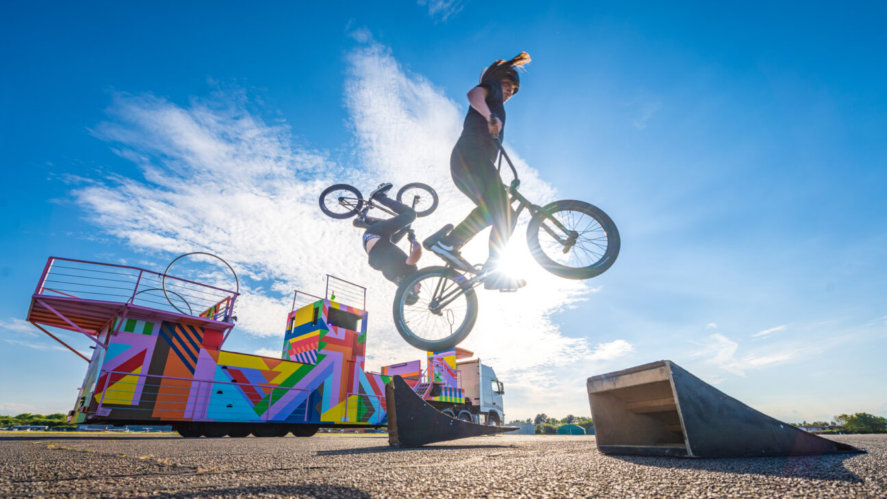 Two stunt bike riders caught mid jump against a sunny backdrop, with our float for the jubilee colourful in the distance