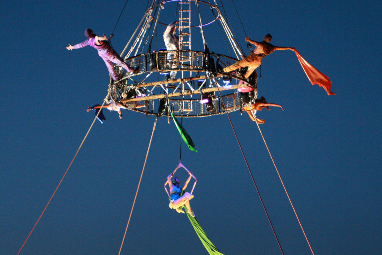 Aerialist perform off our aerial carousel, suspended from a crane. They are dressed colourfully, and perform against a dusk night sky