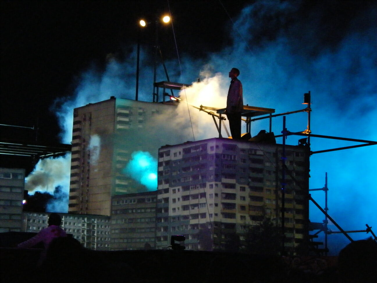 Cirque Bijou, Project 3Sixty. A man is stood on top of a piece of set that looks like a block of flats. There's a dark background with two stage lights drawing attention to smoke between the man and the other blocks of flats, some taller, some shorter.