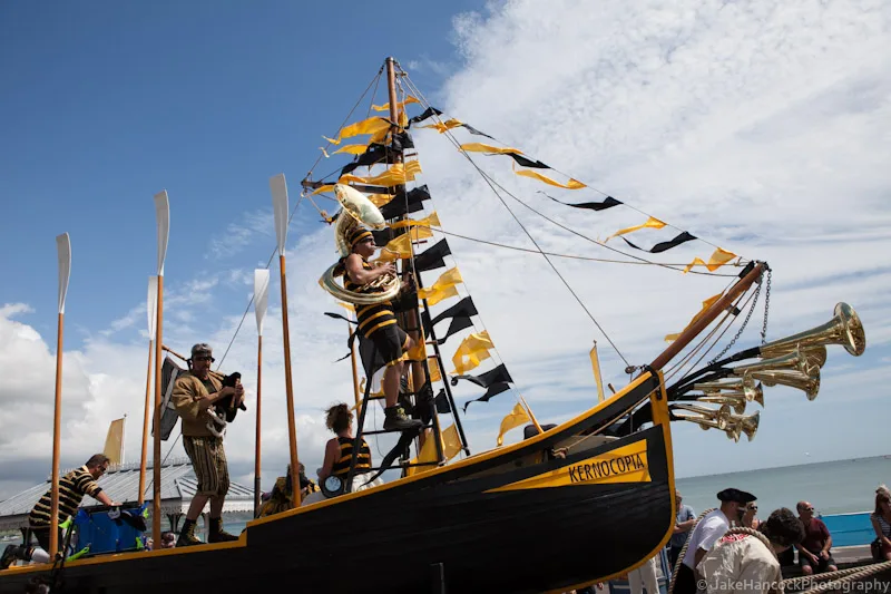 A boat decorated in yellow and black flags with 5 performers in matching costume. One performer is blowing a large horn.