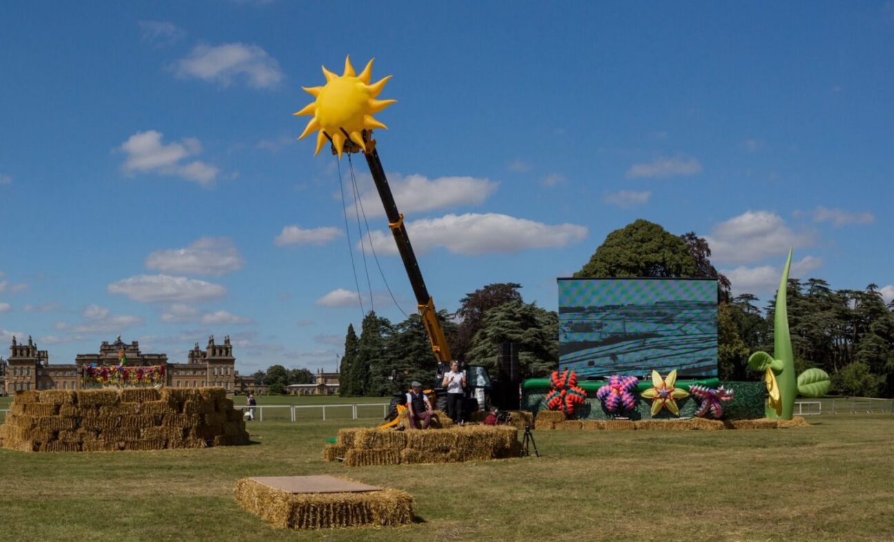 A large inflatable sun sits atop a cherry picker, with a screen in a field surrounded by hay bales