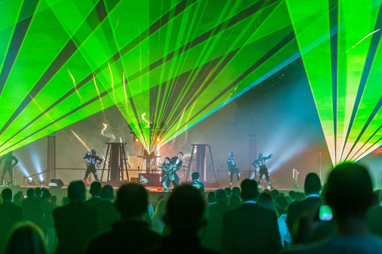 Goodwood, Festival of Speed. The audience, blurred, stand looking at the performers on stage where green crossing beams of light are shooting upwards and outwards. There are 3 dancers in black and neon suits moving. There is a guitarist and another performer centre stage.