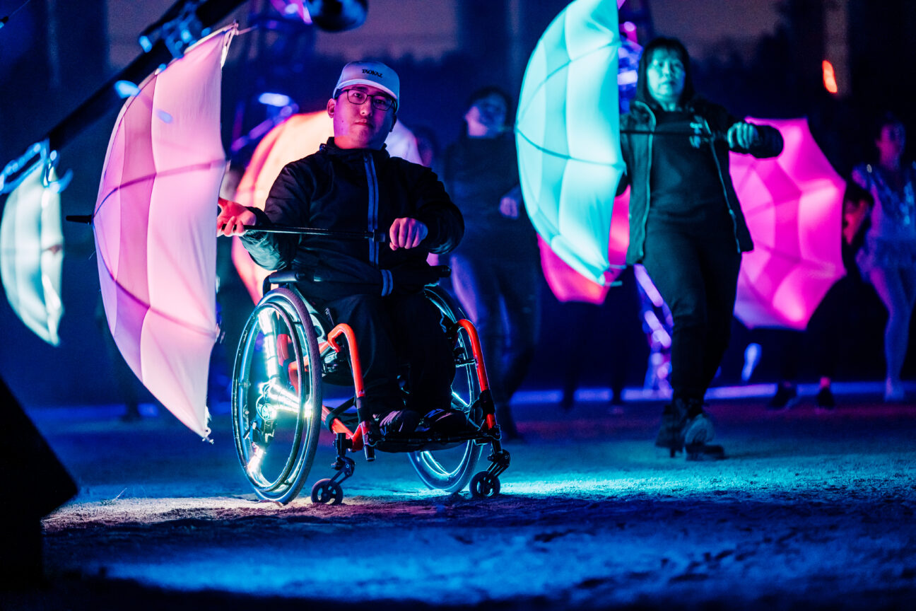 The Umbrella Project. Two performers can be seen, one using a wheelchair, one standing, holding pink and blue LED umbrellas.