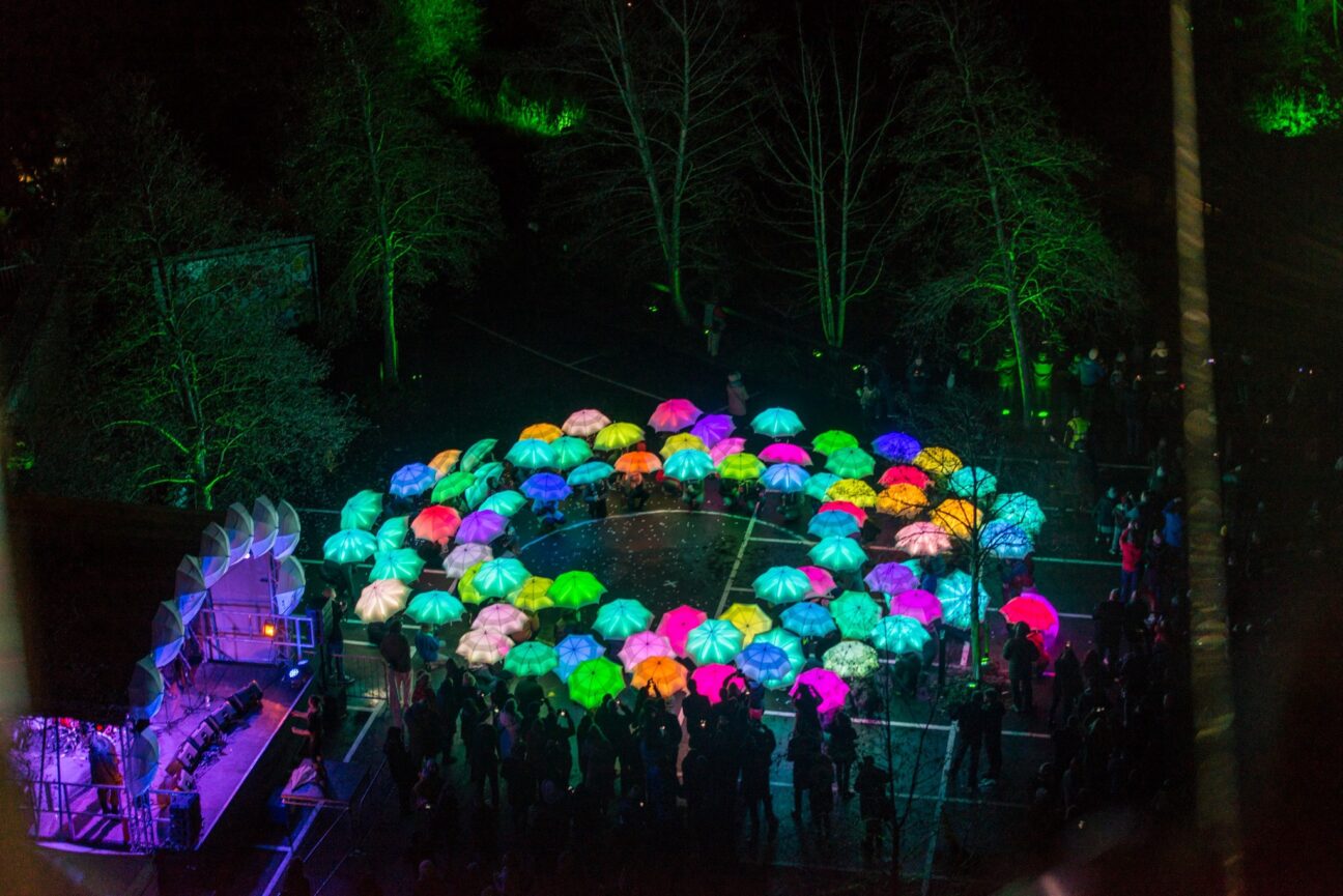 A birds-eye view of a large group of people gathered at night, in a circle. They are all holding coloured LED umbrellas above their heads. They are gathered in Bristol City centre surrounded by trees that are lit by a green light.