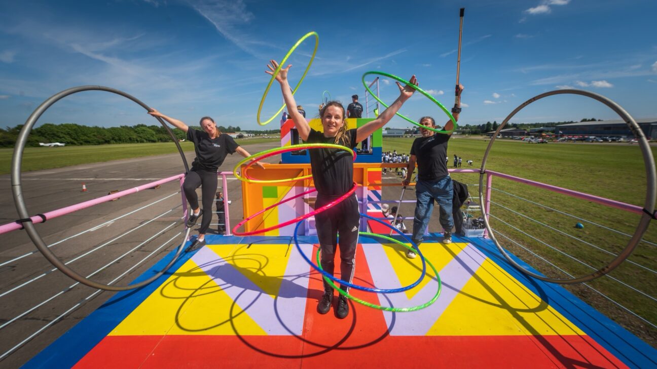 Three performers are stood on a colourful stage float, they are wearing Cirque Bijou black tops, black trousers and jeans. A female performer is stood stage centre circling five hoops around her body and one around each hand. A male performer to the right of her raises his arm and crutch whilst cheering at her. To the left, another female performer is perched on the railings of the float smiling at the central performer as well.