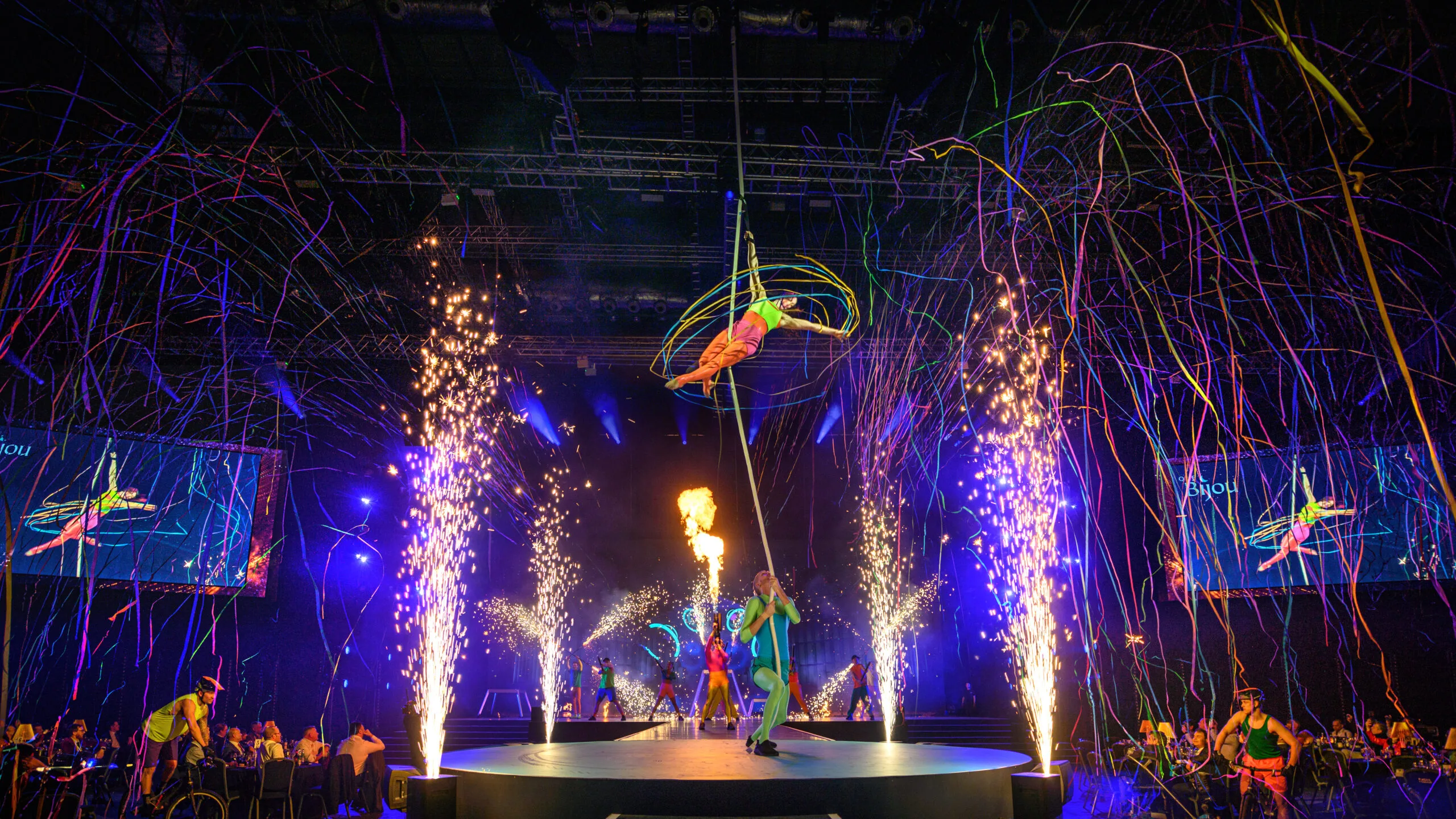 An aerialist hangs from a rope in the centre of the image, surrounded by pyrotechnics going off around her