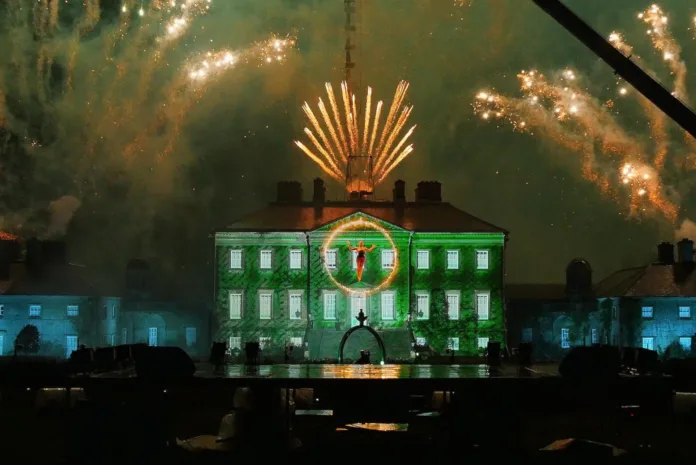 A stately home is illuminated green with projections, with fireworks going off all around it. In the foreground and aerialist performers in a hoop of fire