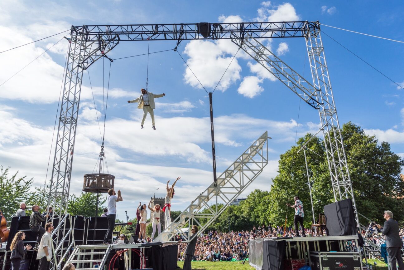 A large truss stands in a field in front of a huge audience, with an aerialist suspended in the air