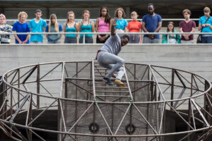 Extraordinary Bodies performer David Ellington swings from metal structure in What Am I Worth? National Theatre River Stage. Image Rob Harris.