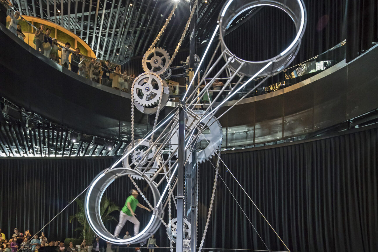 Wheel of Energy structure. Two large metal wheels and 6 metal cogs in the background. There is a performer in the bottom left wheel moving it round.