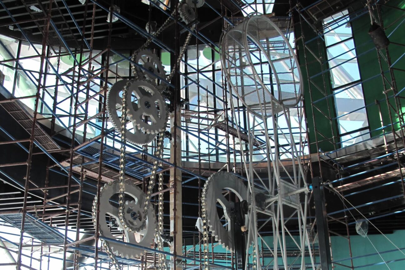 The Wheel of Energy set in Kazakhstan. A large metal wheel centre right, suspended in the air on a metal structure with 5 large grey metal cogs behind it, surrounded by scaffolding.