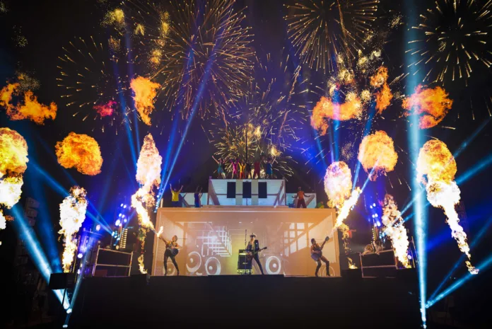 On a large stage, there are 3 performers on the bottom level all playing guitar. Flames shoot out of the two female guitarists guitars either side. There are flames and fireworks all around the stage. Performers on the above two levels are stood proudly with there arms up above them in a 'Y' shape.