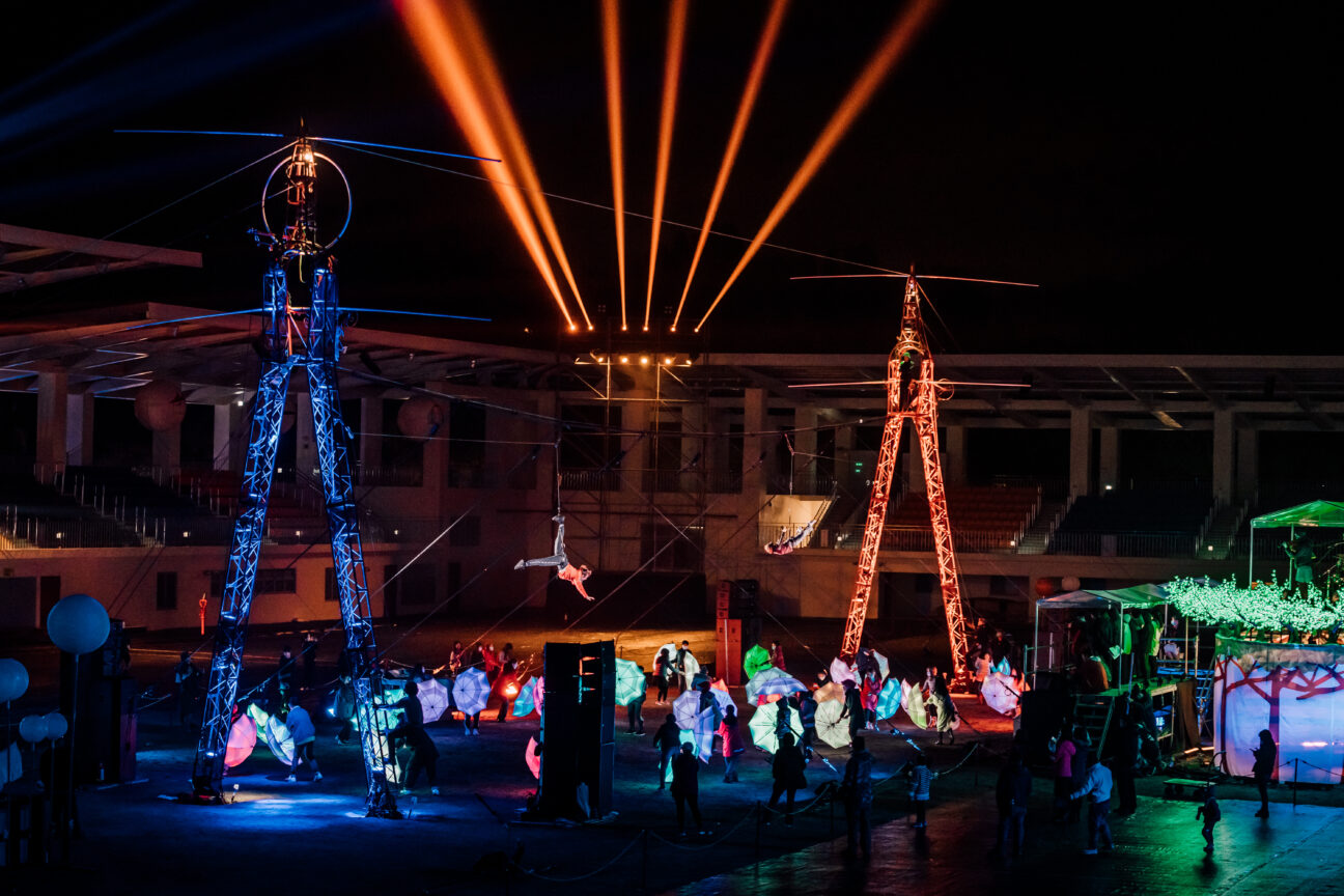 A shot of the high wire, illuminated with lights. An aerialist dangles and performs from the wire, whilst a dance with the LED umbrellas happens below her