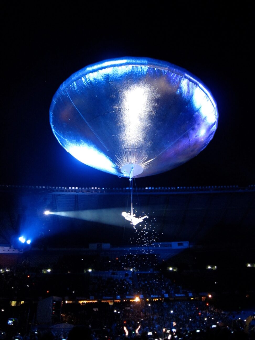 A performer is suspended from a helium filled UFO-shaped balloon. A bright, white light shines on the performer and the audience are in darkness.